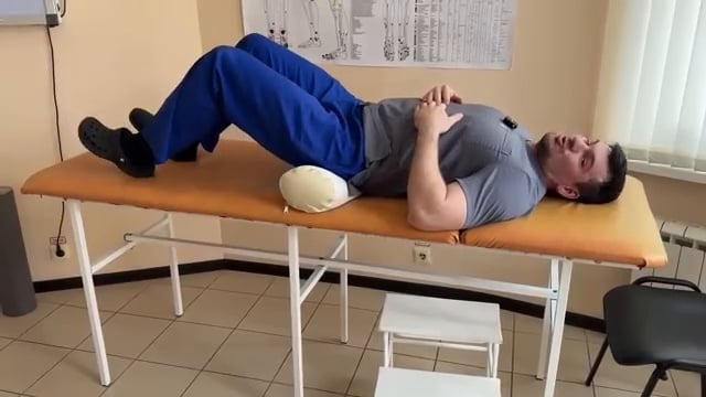 Le thérapeute ajuste la position des jambes (genoux pliés) pour maximiser la tension abdominale et le contact du bas du dos avec la surface.